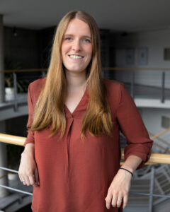 Auf dem Bild ist Katharina Schiffmann zu sehen. Sie trägt eine lockere Bluse, lehnt an einem Geländer und schaut in die Kamera.