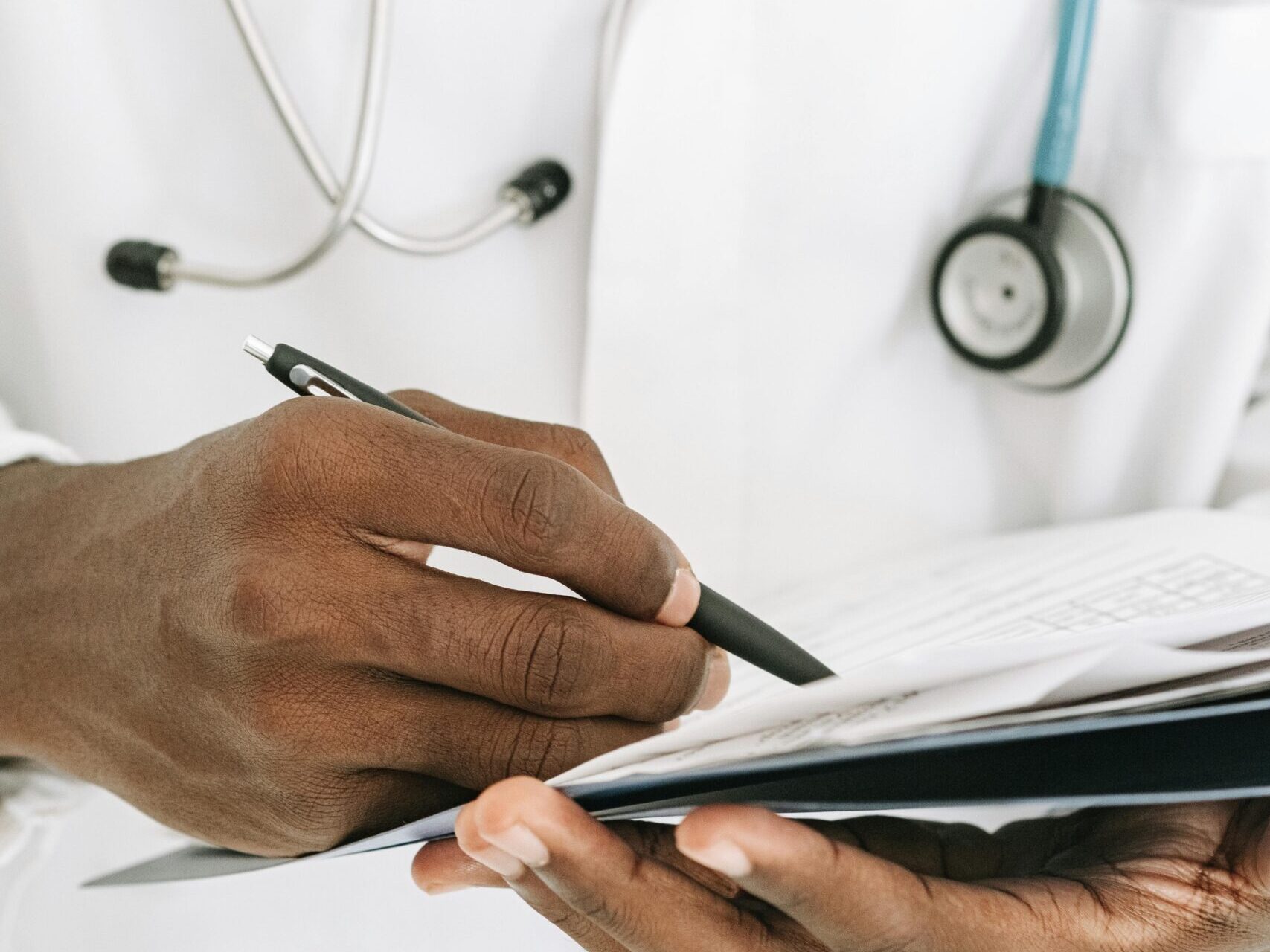 Ein Arzt füllt mit der rechten Hand Dokumente aus, die in seiner linken Hand liegen.