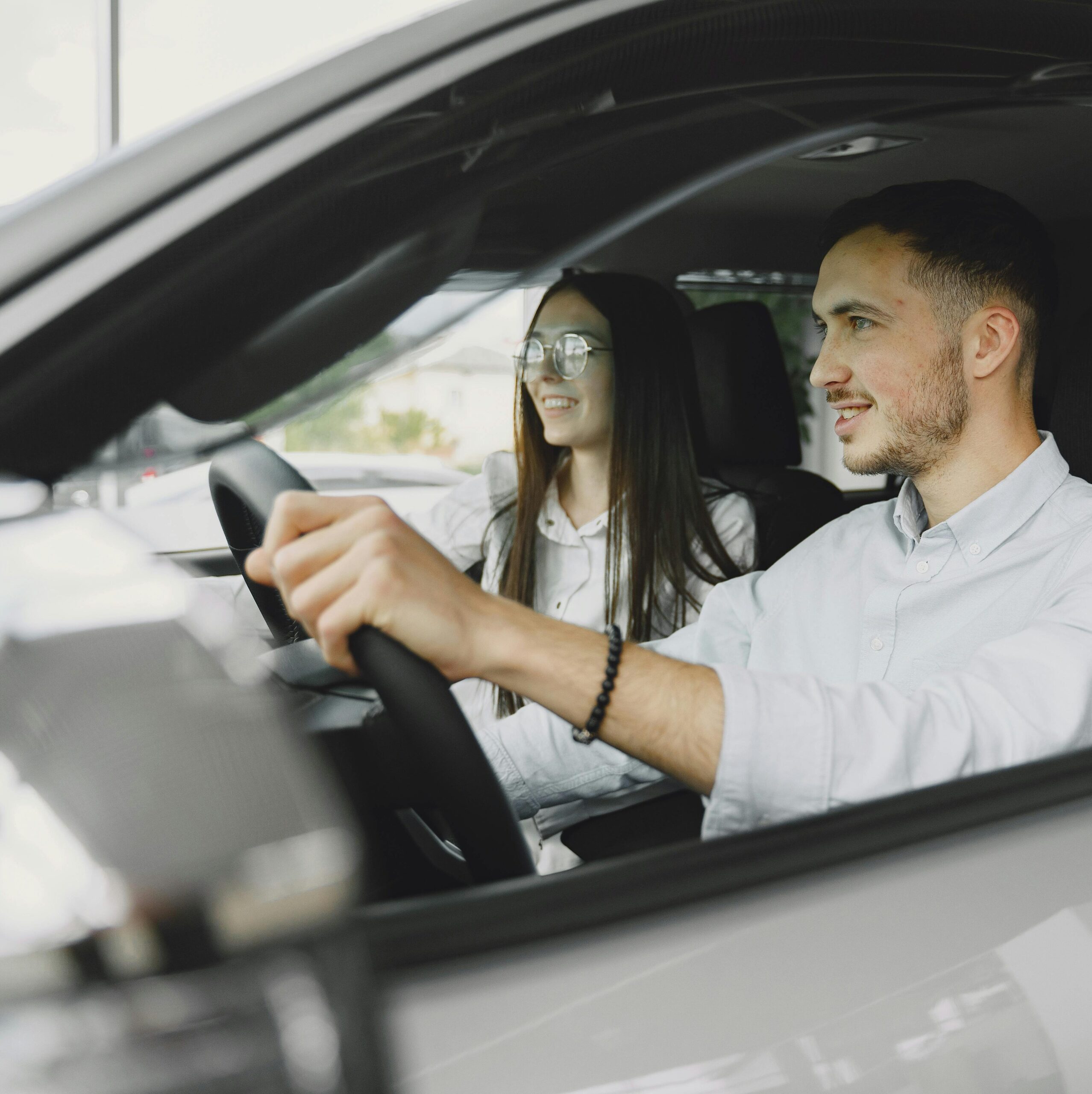 Zwei Personen sitzen in einem Auto; eine Person am Steuer, die andere auf dem Beifahrersitz. Beide blicken nach vorne.
