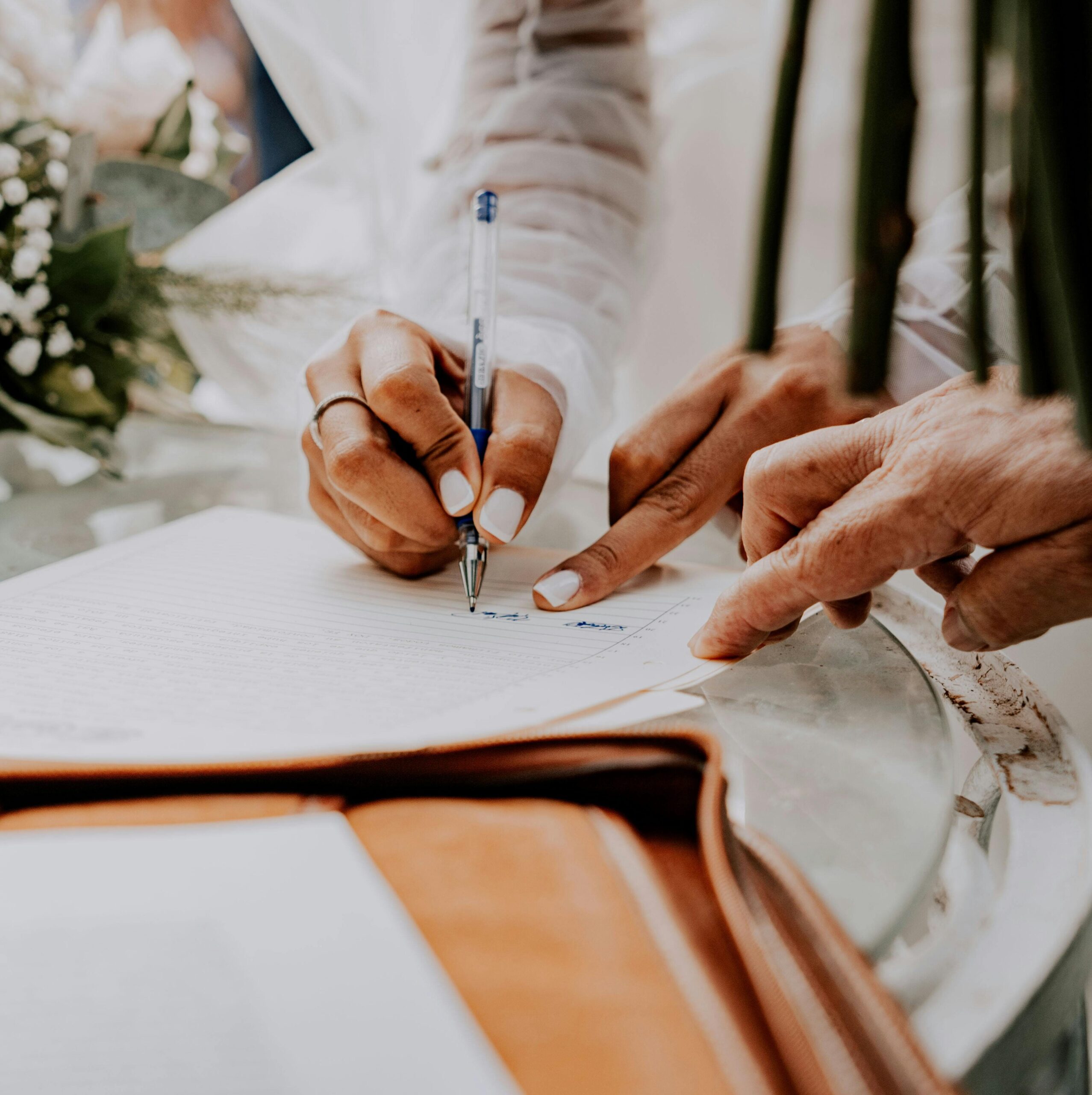 Eine Person unterschreibt ein Dokument mit einem Stift, während eine zweite Hand auf die Unterschriftsstelle zeigt; daneben liegt ein Blumenstrauß.